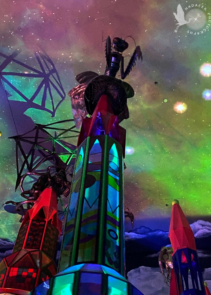 Colorful spires reach up to a space-like sky. Atop the nearest spire is a statue of a praying mantis.